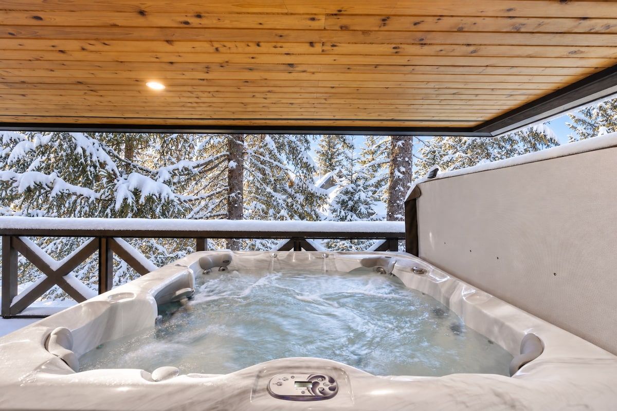 A hot tub with a control panel, surrounded by snow-covered trees and a wooden deck.