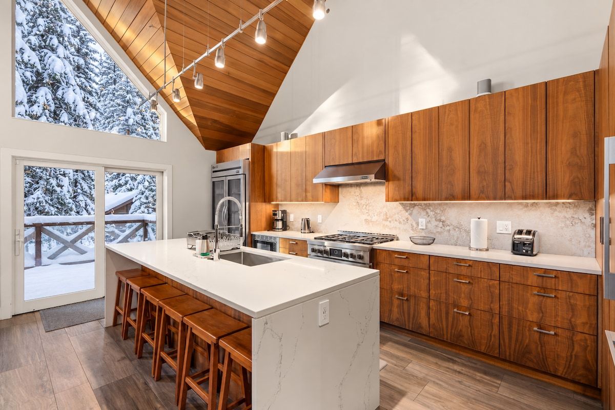 Une cuisine moderne avec des meubles en bois, un grand îlot central doté de tabourets de bar et une vue sur les arbres enneigés par les fenêtres.
