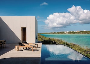 A modern outdoor pool area with a view of the ocean and a building with a door and seating. - Secluded Oceanfront Villa | Direct Water Access (Providenciales)