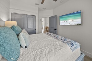 Quarto com cama, travesseiros, mesa de cabeceira, abajur e TV na parede exibindo uma cena de praia.