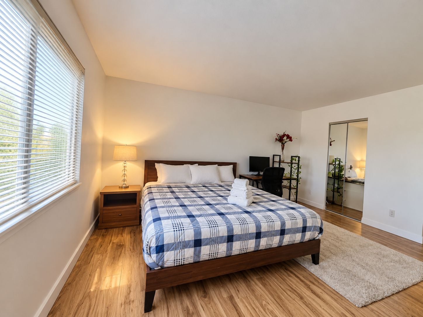 A bedroom with a wooden bed, a nightstand, a lamp, a desk with a computer, and a sliding door closet.