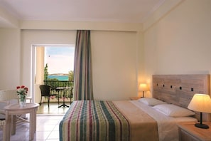 A hotel room with a bed, bedside lamps, a small table with a vase of flowers, a chair, and a balcony with a view of the sea. - Portes Beach Hotel (Nea Propontida)