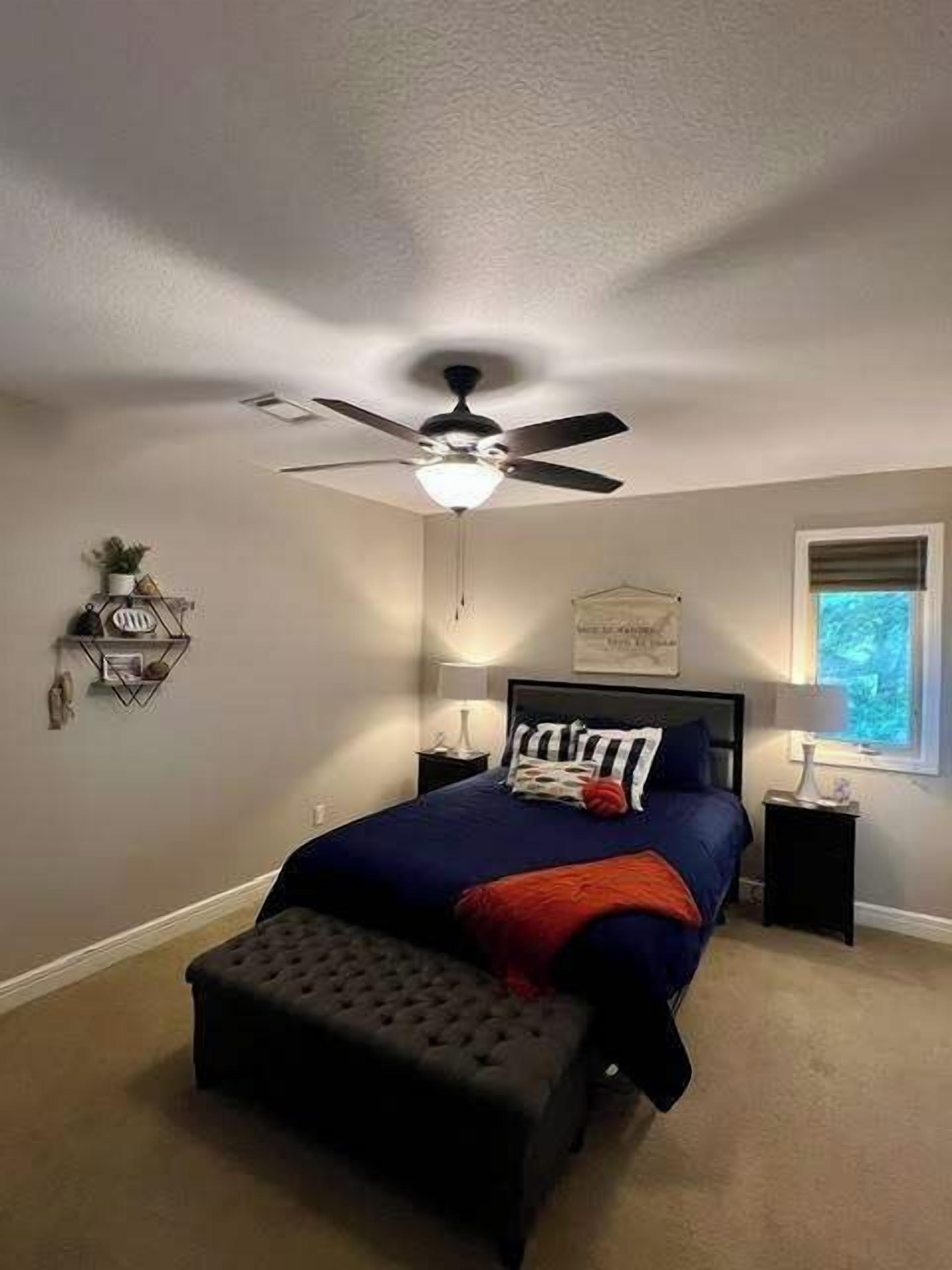 Un dormitorio con cama, ventilador de techo, mesitas de noche, ventana y estante en la pared.