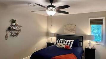 Un dormitorio con cama, ventilador de techo, mesitas de noche, ventana y estante en la pared.