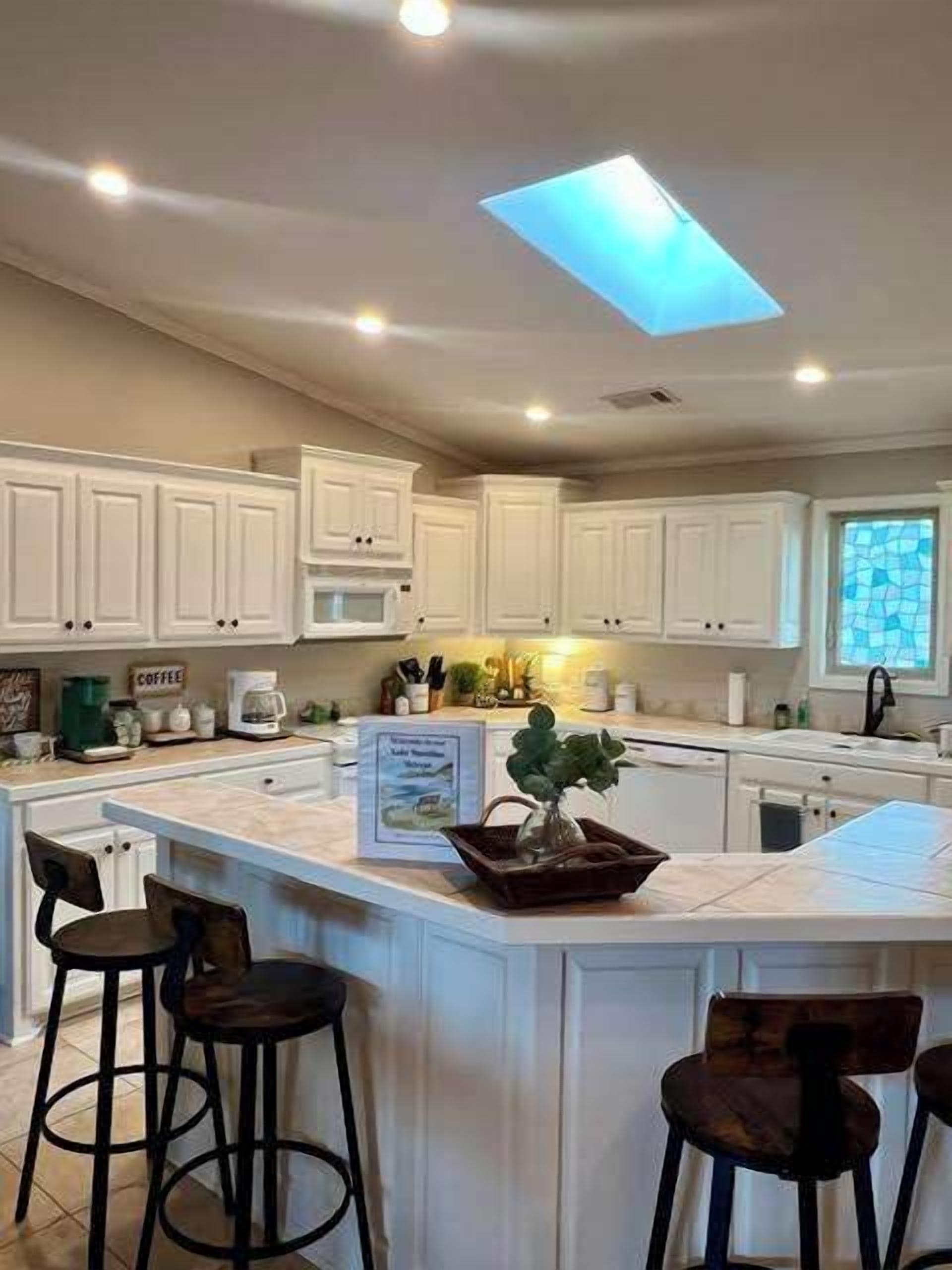 A kitchen with white cabinets, a central island with stools, a skylight, and a window with a view.