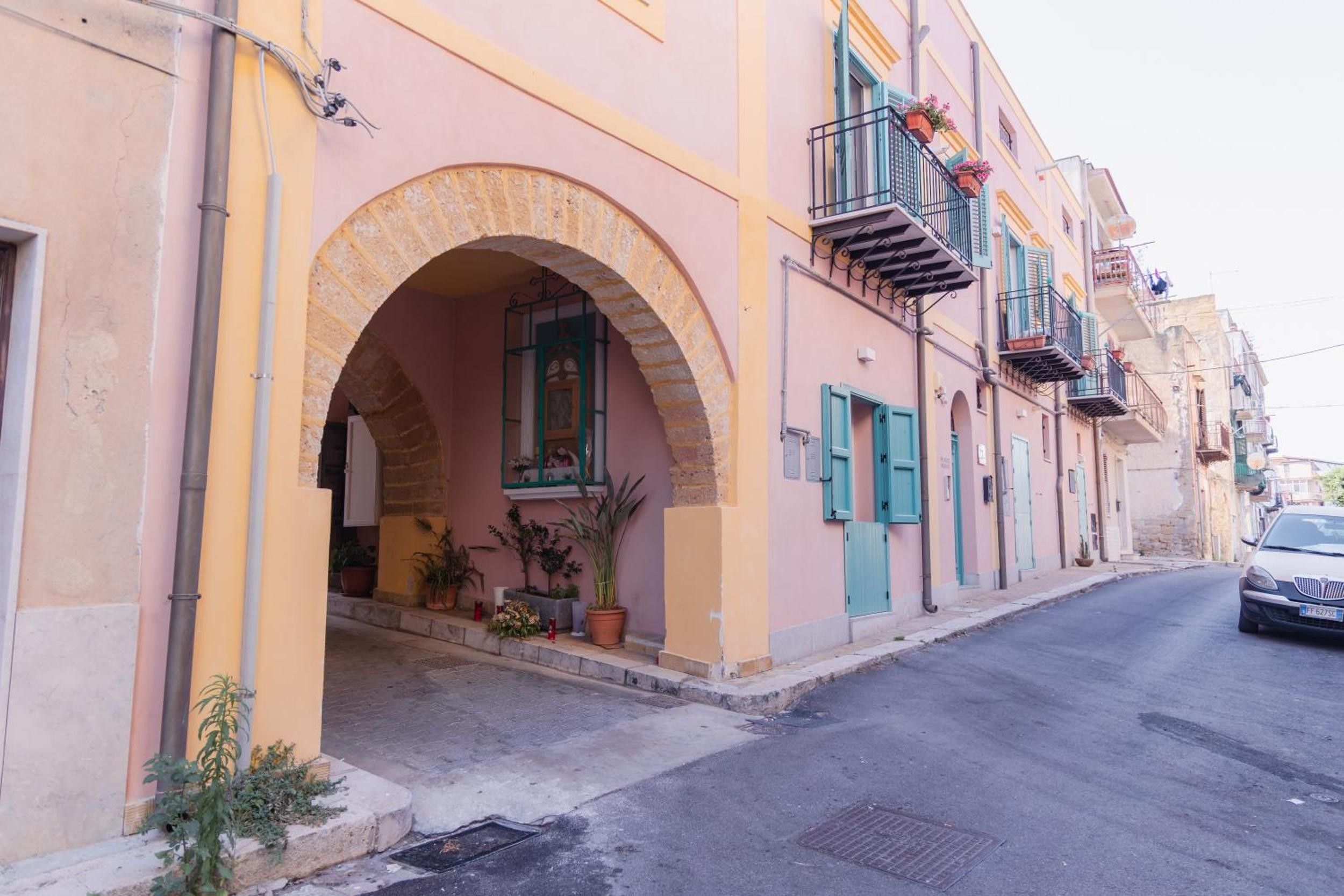 A street with a building featuring an arched entrance, potted plants, and a vehicle parked on the side.