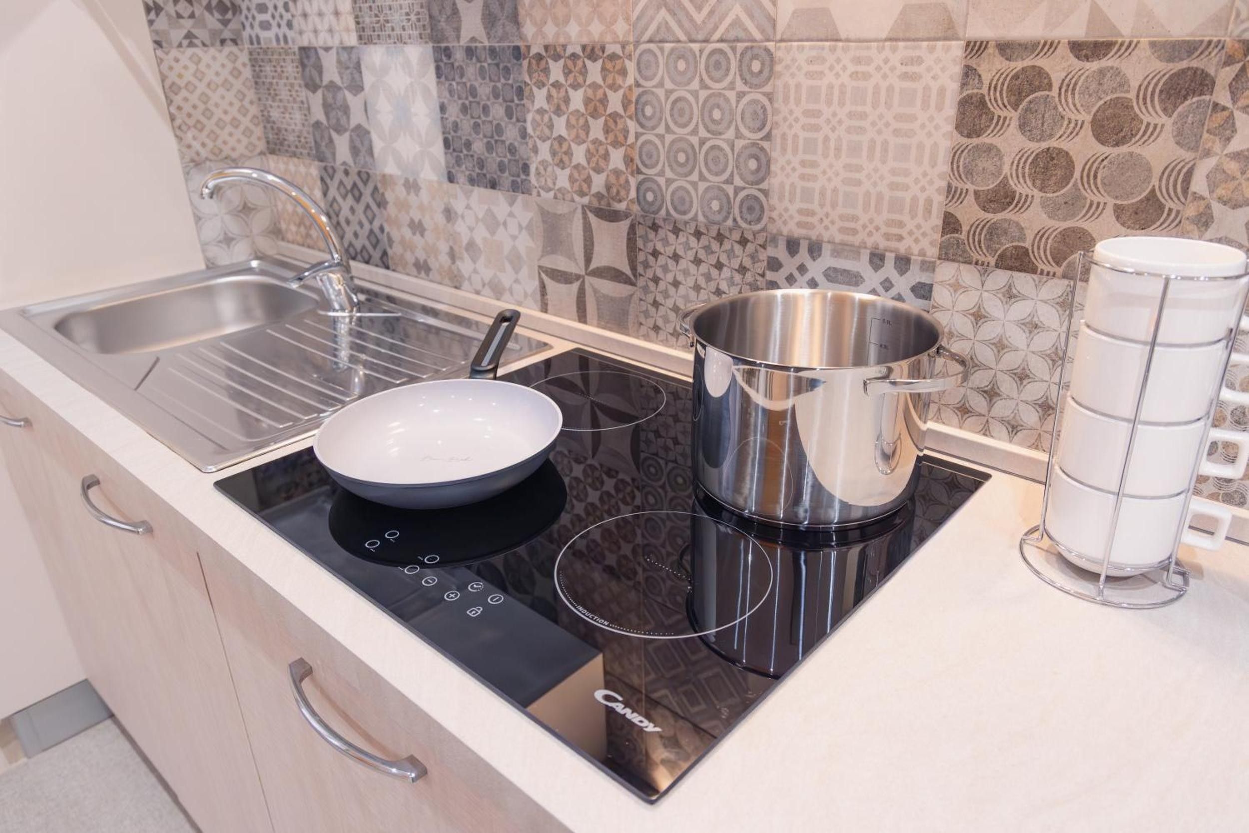 Una cocina moderna con una placa de inducción negra, una olla de acero inoxidable, una sartén blanca y una taza blanca sobre una encimera blanca.