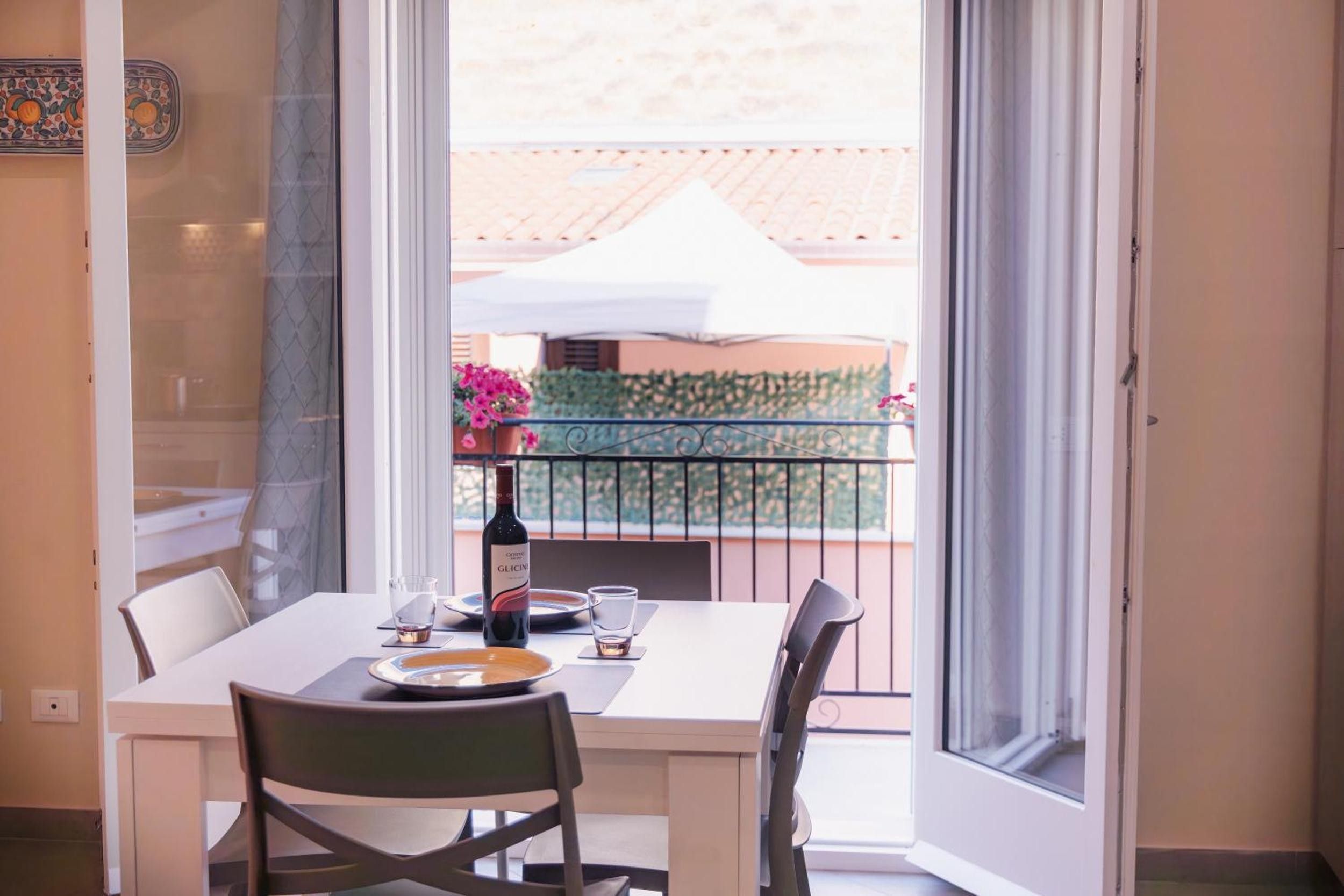 A dining area with a table set for two, a bottle of wine, and a view of a balcony with a white umbrella and greenery.