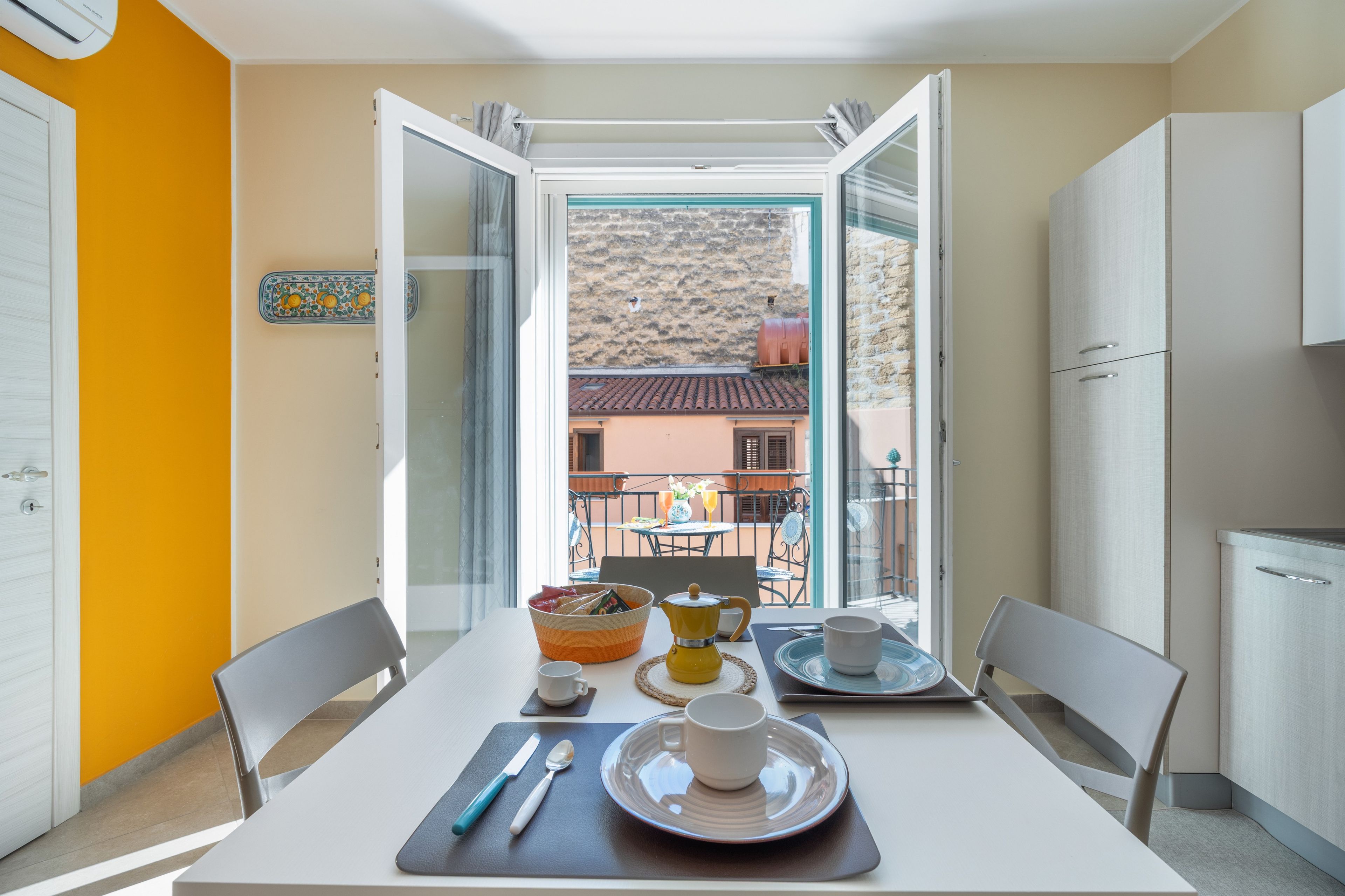 Un comedor con una mesa lista para dos, una ventana con puerta abierta y vista a un edificio con tejado de tejas.