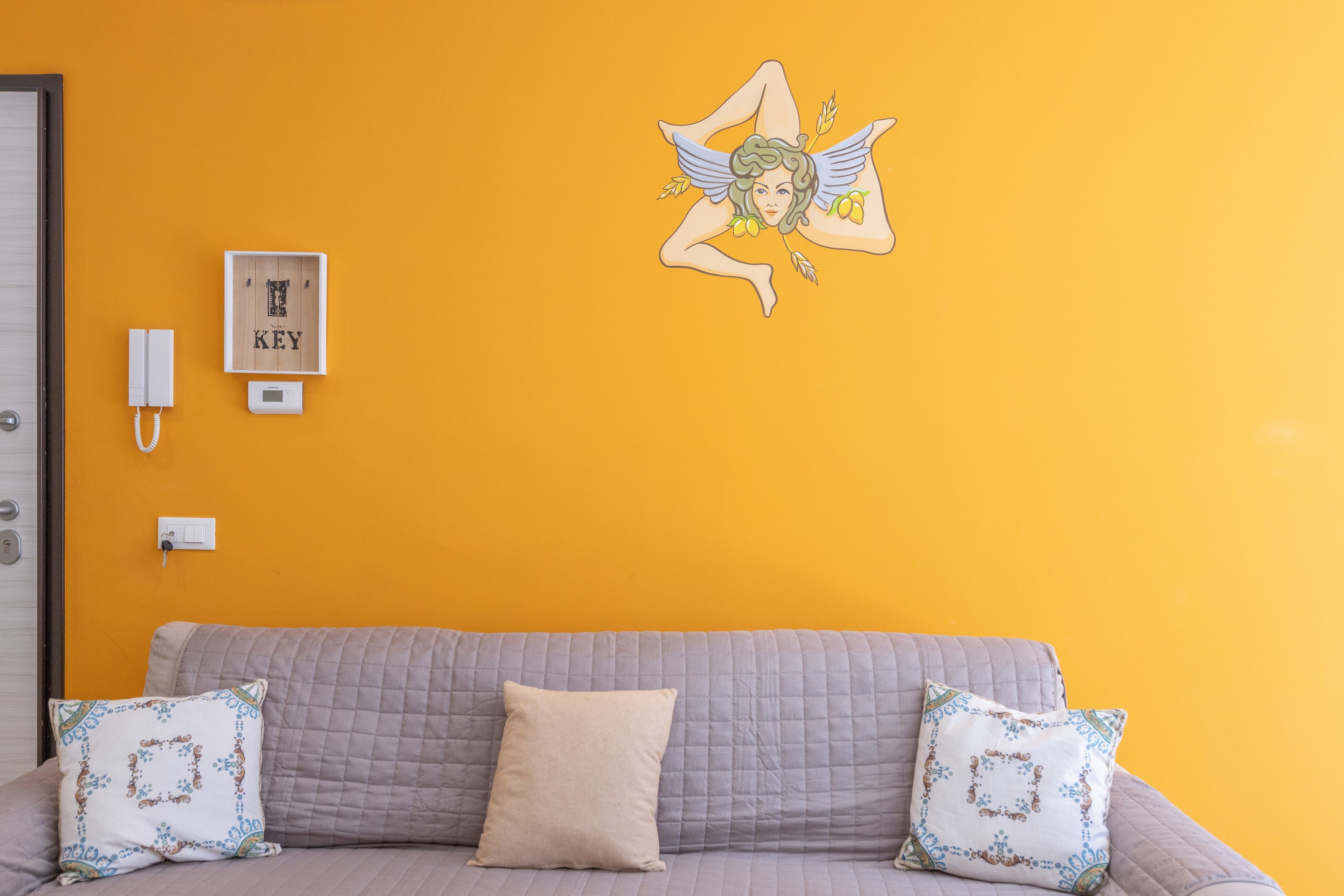 A room with a yellow wall, a white door, a grey sofa with two patterned pillows, a framed artwork, a white telephone, and a thermostat.