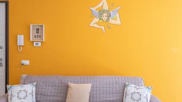 A room with a yellow wall, a white door, a grey sofa with two patterned pillows, a framed artwork, a white telephone, and a thermostat.