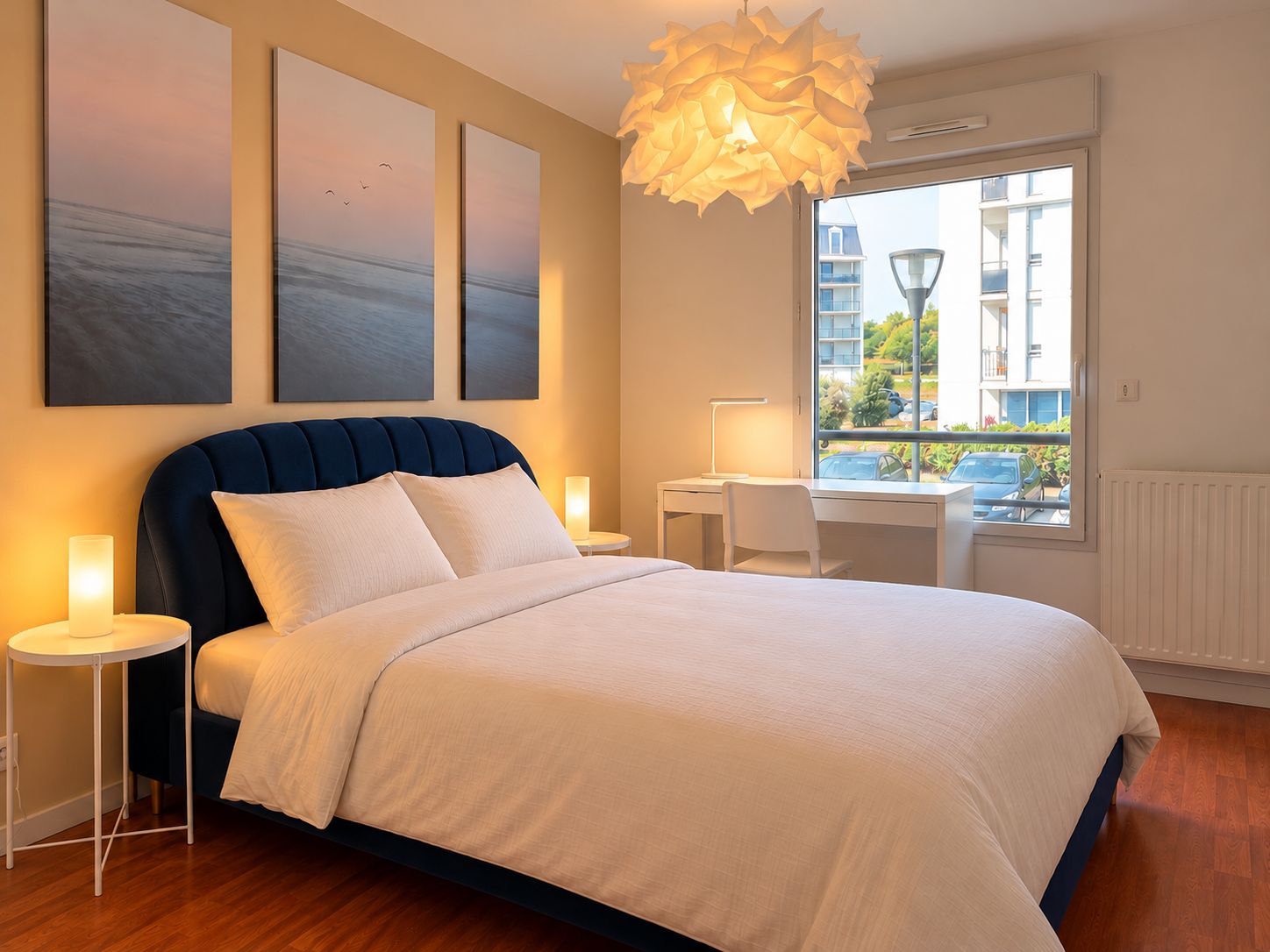Ein Schlafzimmer mit einem Bett, Nachttischen, einem Schreibtisch und einem Fenster mit Blick auf Gebäude und Grünflächen.
