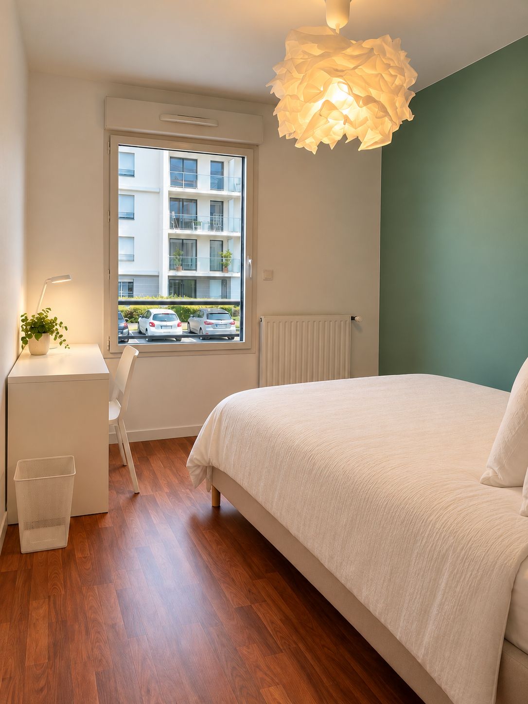 Ein Schlafzimmer mit einem großen Bett, einem Fenster mit Blick auf Gebäude, einem Schreibtisch mit Lampe und einer Heizung.