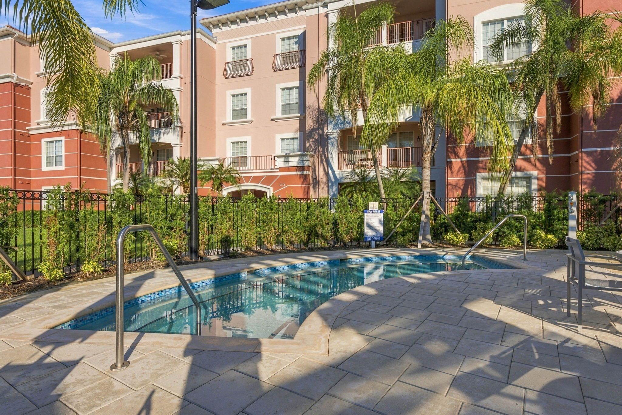 A swimming pool with a tiled deck, surrounded by a fence and palm trees.
