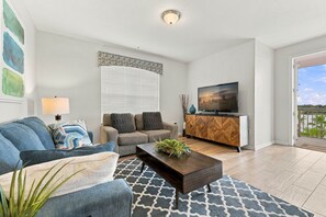 A modern living room with a sofa, a coffee table, a TV, and a balcony. - Amazing 4BR at Vista Cay With Gorgeous Lake View (Orlando)