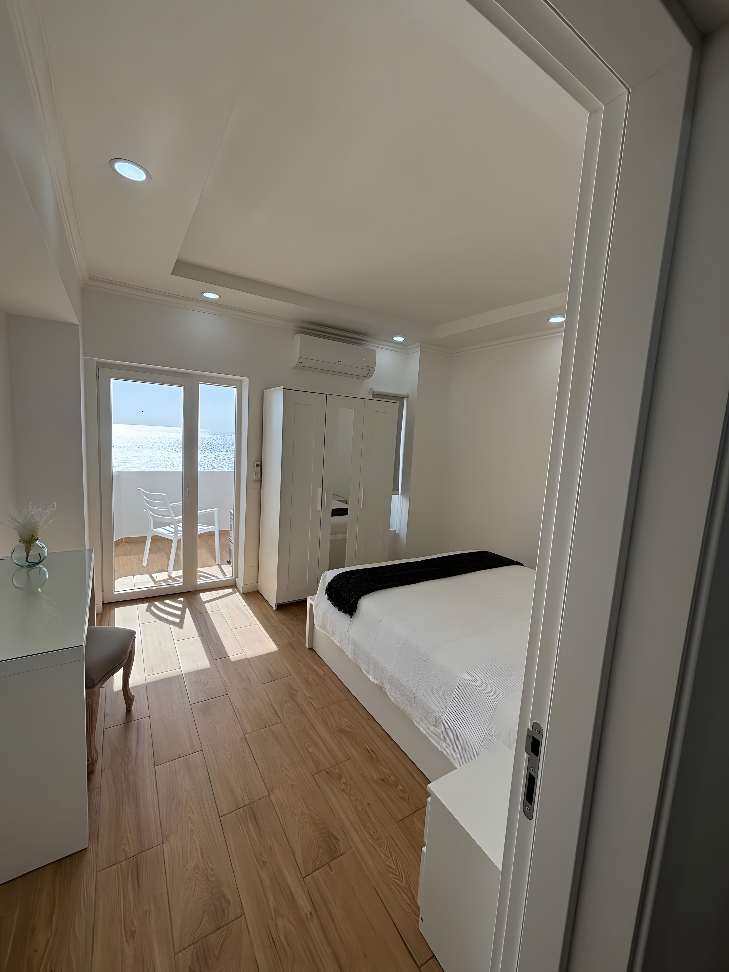 A bedroom with a bed, a desk, and a chair. There is a sliding door leading to a balcony with a view of the sea.