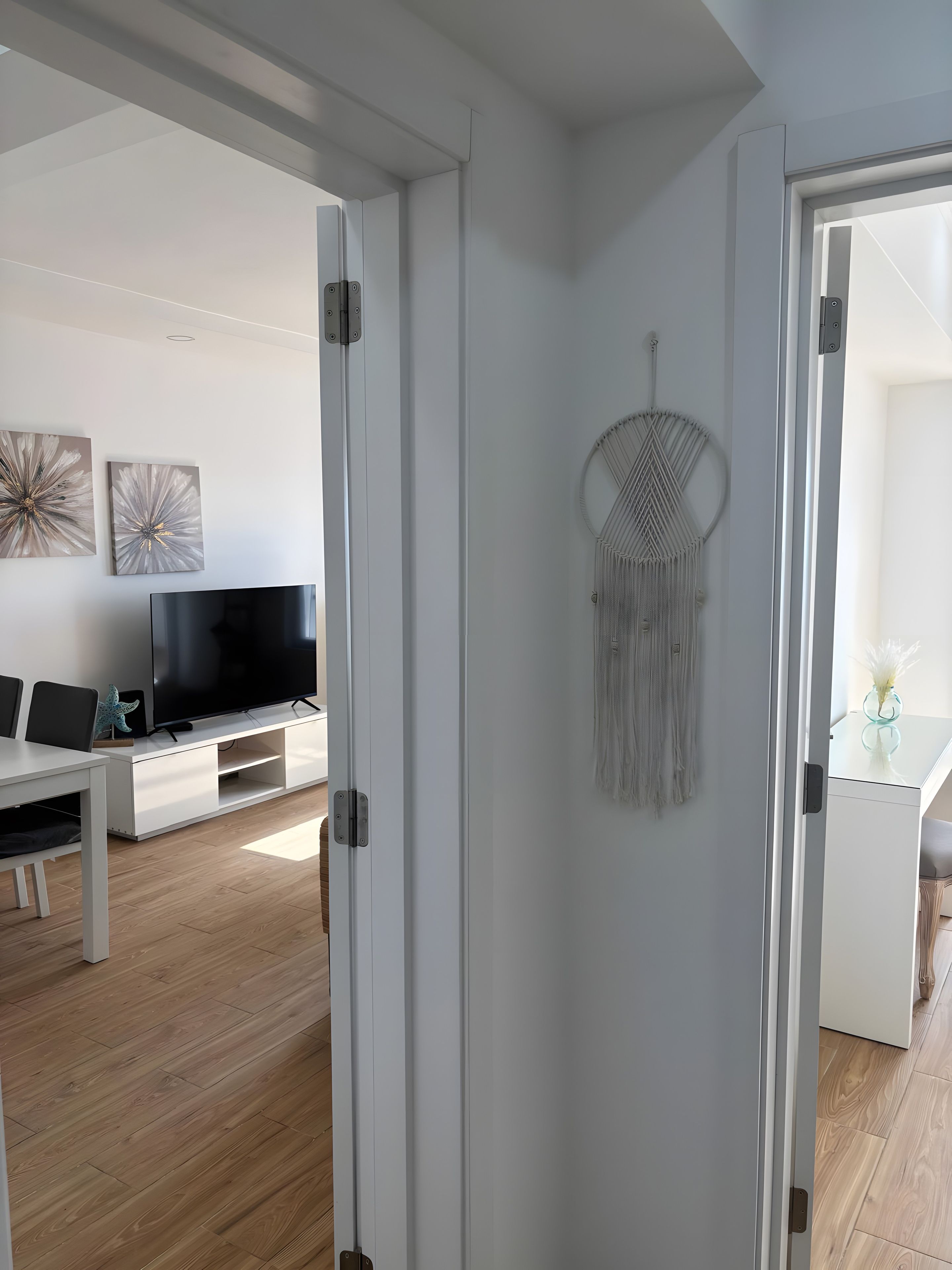 Een interieur met een witte deur, een decoratief object aan de muur, een flatscreen televisie en een eethoek met een tafel en stoelen.