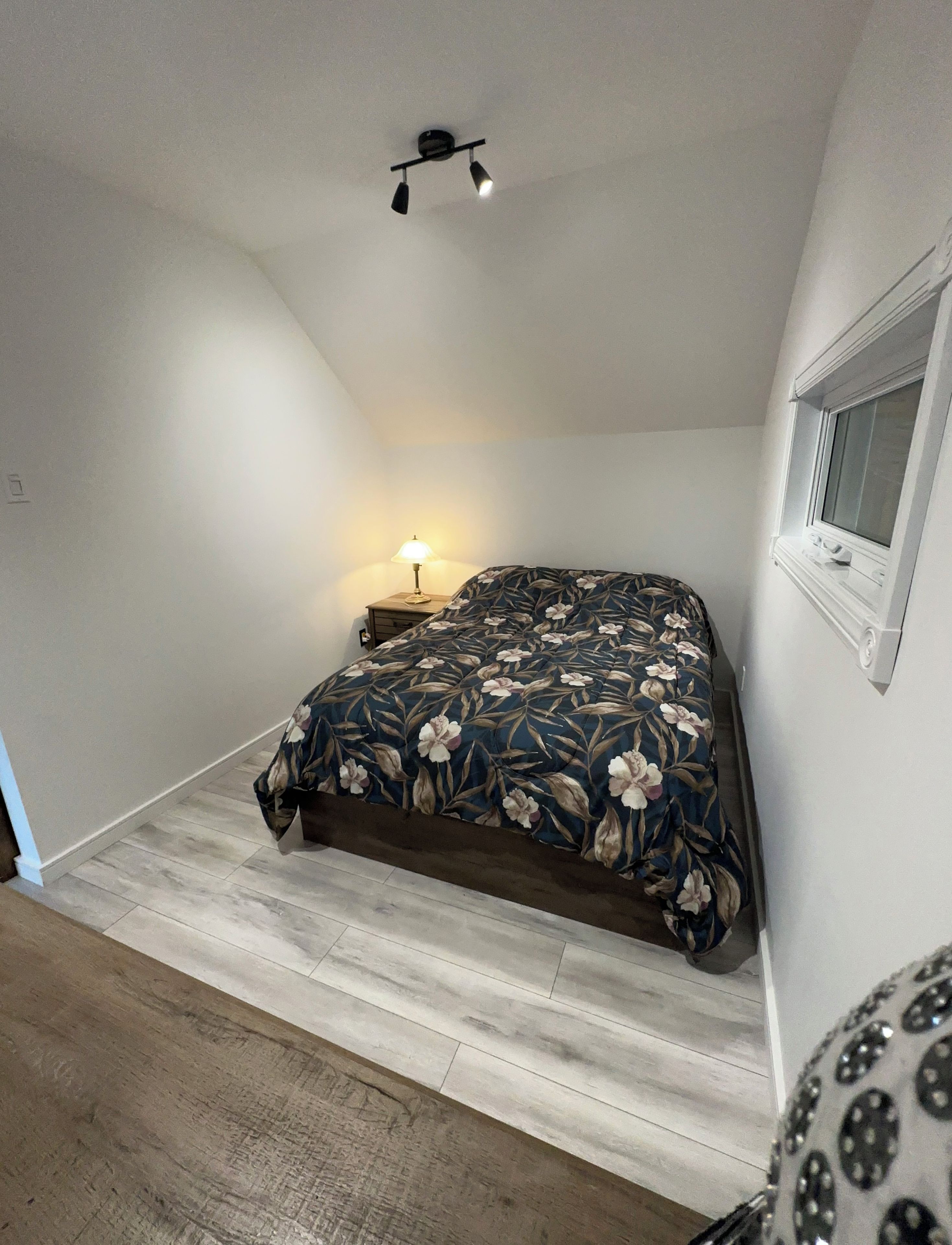 Une chambre à coucher comprenant un lit, une table de chevet, une lampe, un luminaire au plafond et une fenêtre.