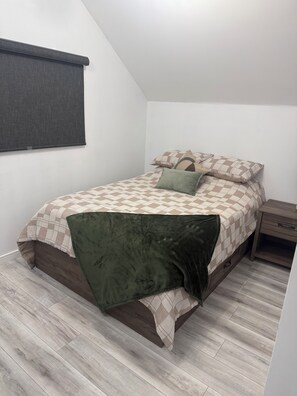 Ein Schlafzimmer mit einem Bett, einem Nachttisch, einem Fenster mit Jalousie und einem Holzboden.