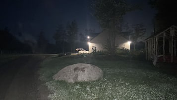 A nighttime outdoor scene with a large rock in the foreground, a white car parked under a tree, and a building with lights on in the background.