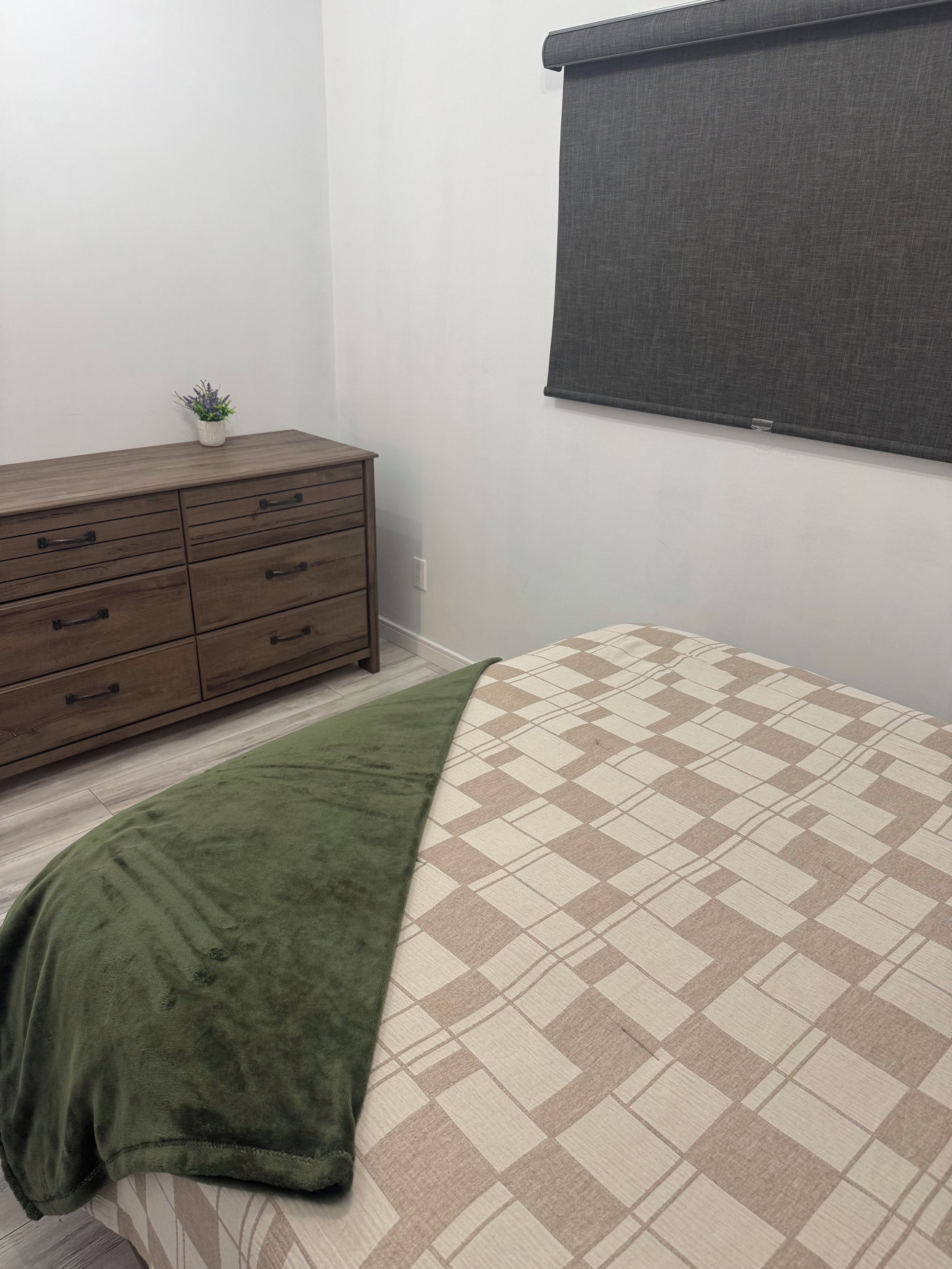 A bedroom with a bed, a dresser, a small plant, and a window with a blind.