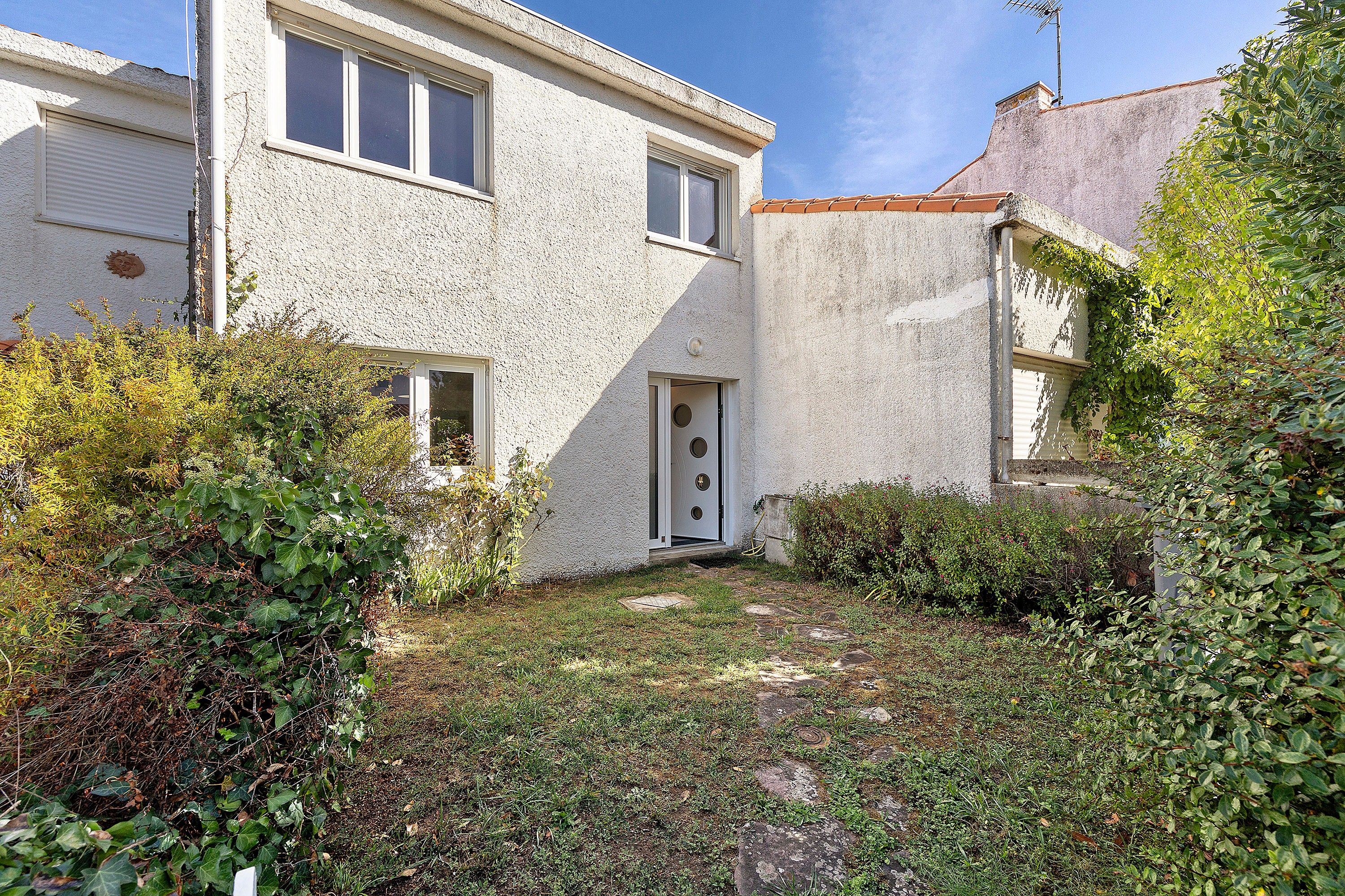 Un bâtiment à deux étages, à l’extérieur blanc, avec une porte dotée d’une fenêtre circulaire et un petit jardin agrémenté de diverses plantes et d’un sentier en pierre.