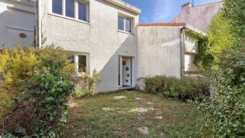 Un bâtiment à deux étages, à l’extérieur blanc, avec une porte dotée d’une fenêtre circulaire et un petit jardin agrémenté de diverses plantes et d’un sentier en pierre.