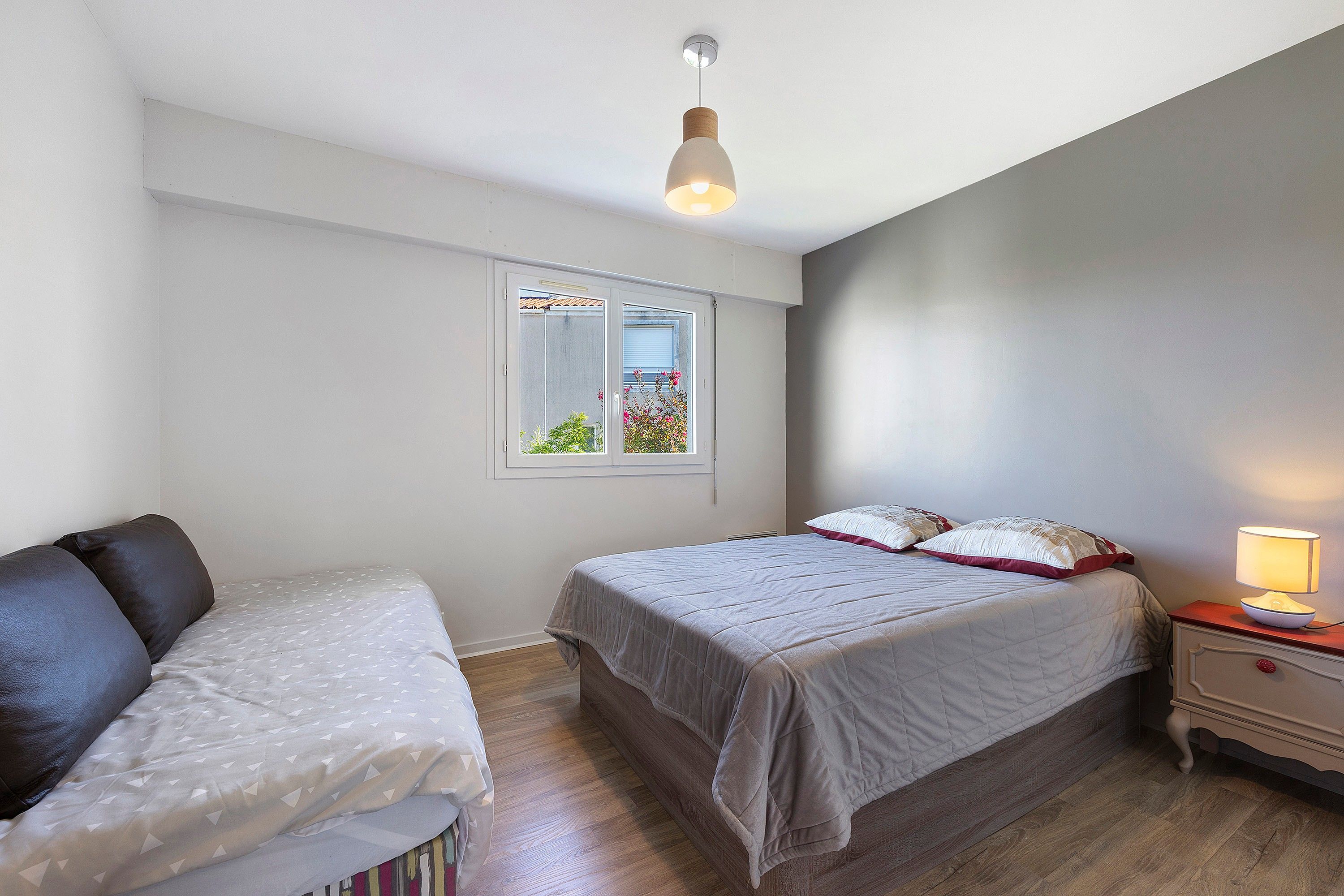 Un dormitorio con cama, mesita de noche con lámpara, ventana con vistas a zonas verdes e iluminación en el techo.