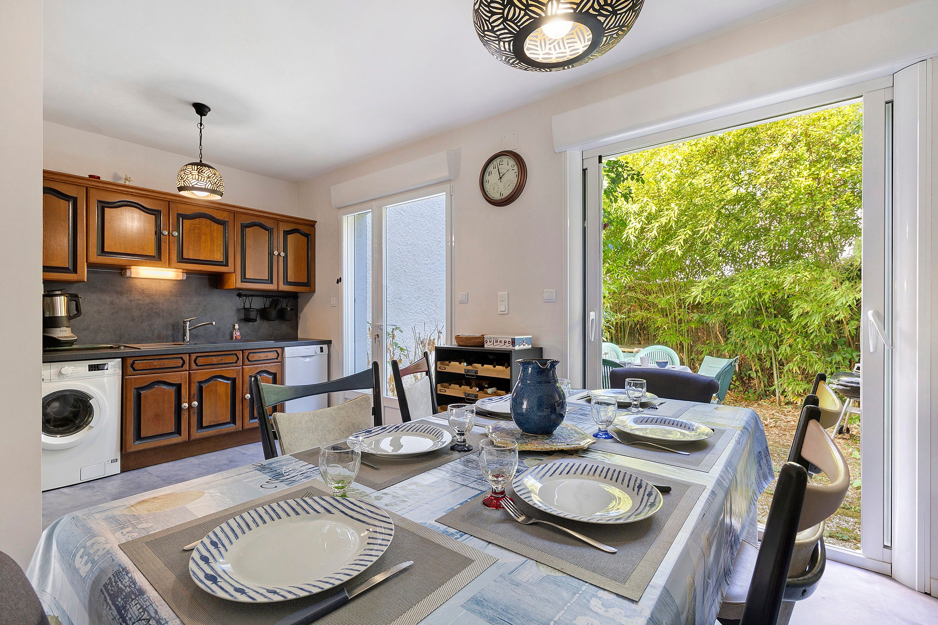 A dining area with a table set for four, a kitchen with wooden cabinets, and a view of a lush garden through a sliding glass door.