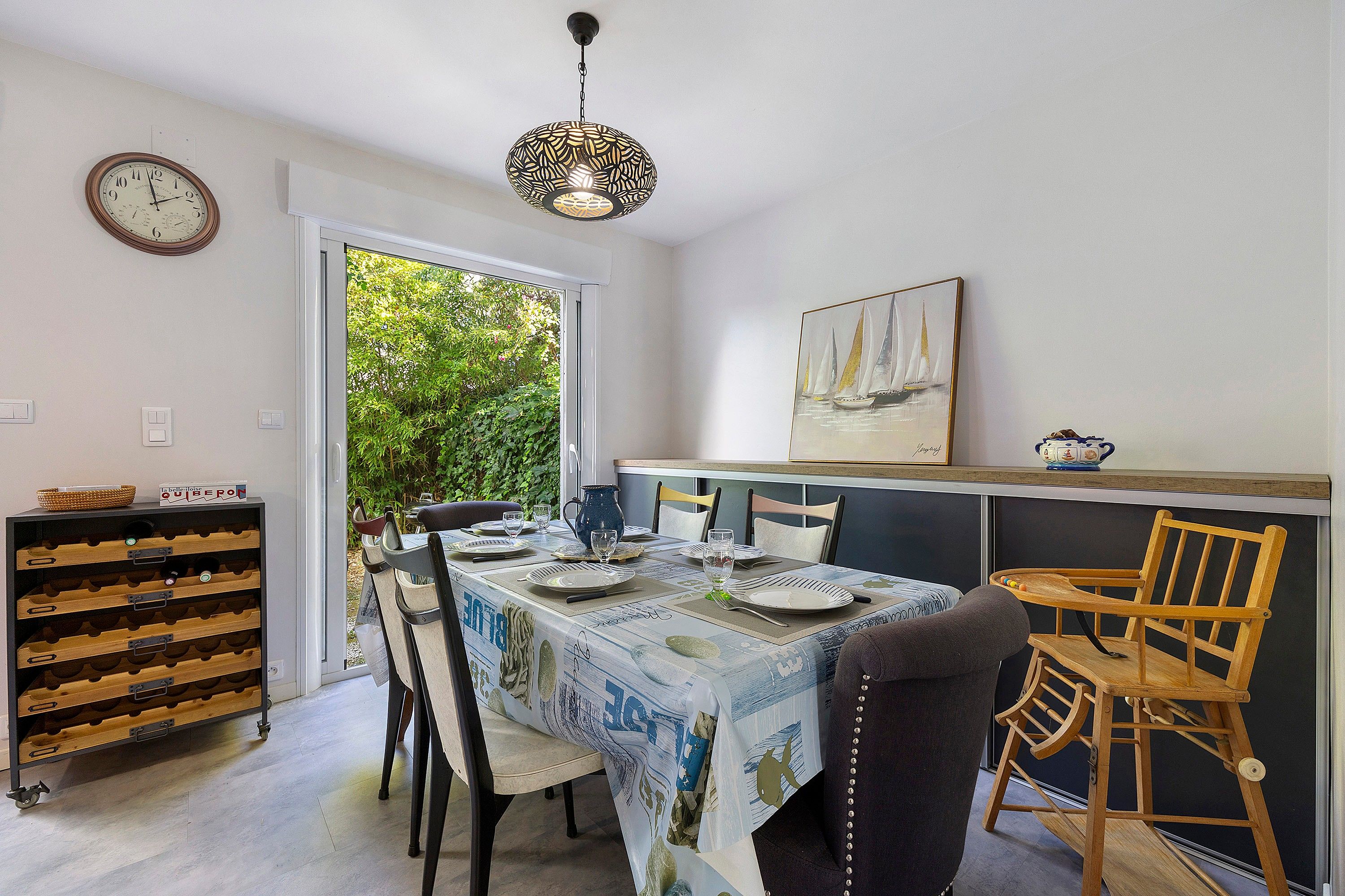 A dining area with a table set for four, a clock on the wall, and a view of greenery outside.