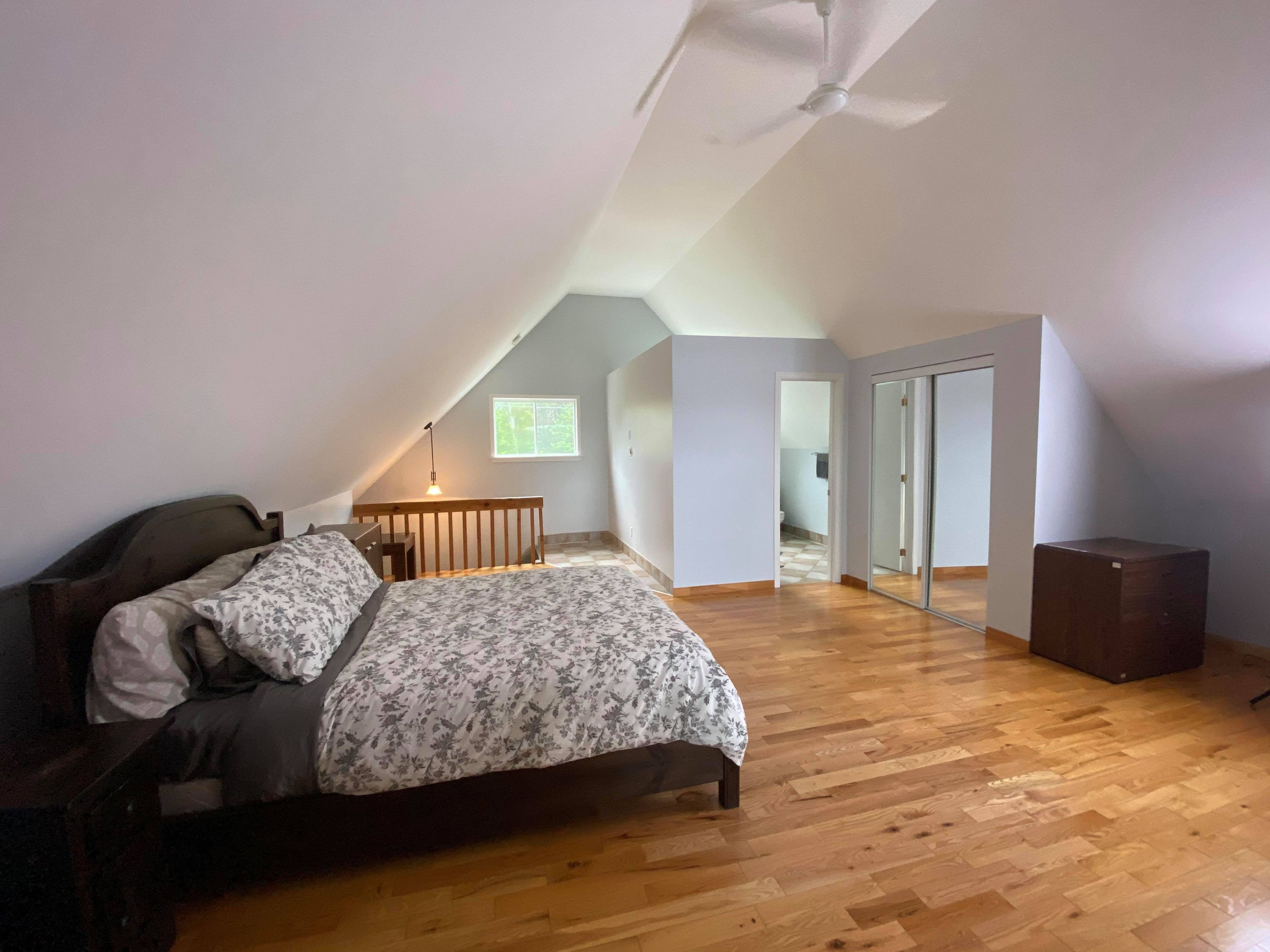 Una camera da letto con soffitto spiovente, un letto, un comodino, una lampada e una finestra.