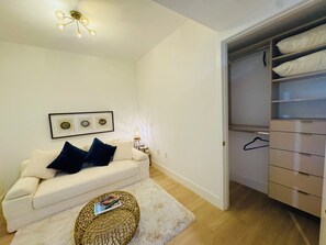 Quarto com sofá branco, um banquinho redondo de fibra natural, quadro pendurado na parede e armário com roupas.