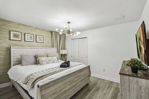 Une chambre à coucher comprenant un lit, une lampe de chevet, un luminaire au plafond, une porte et un téléviseur posé sur un meuble.