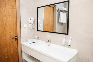 Casa de banho de hotel com lavatório branco, espelho e secador de cabelo.