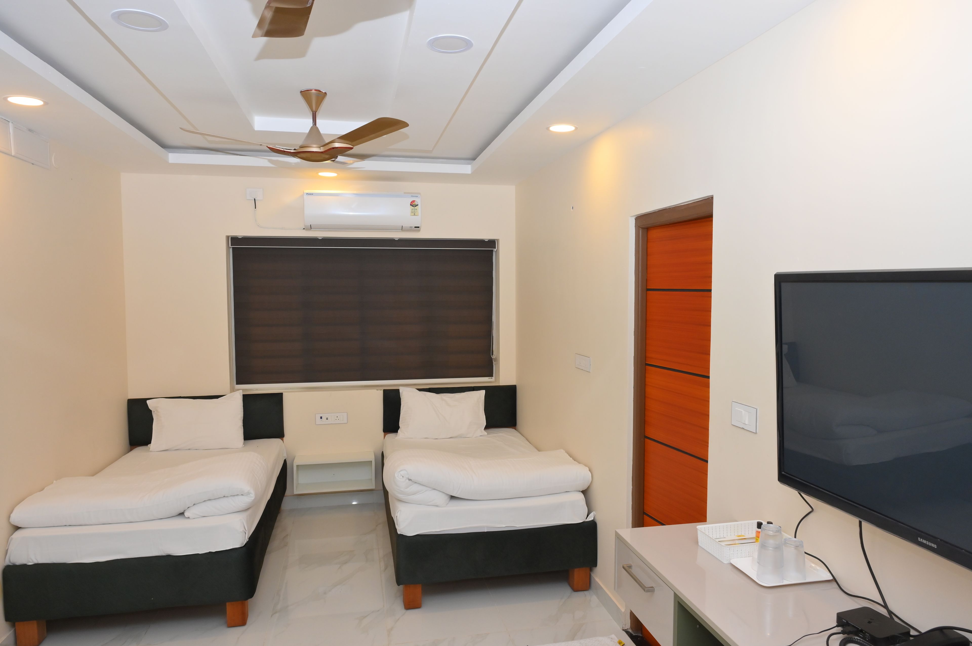 Habitación de hotel con dos camas, ventilador de techo, aire acondicionado, televisión y puerta de madera.