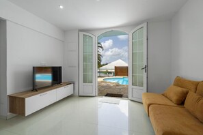 A modern living room with a flat-screen TV, a sofa, and a view of a swimming pool through glass doors. - Appartement Central View (Ducos)
