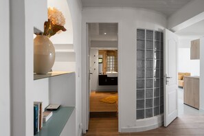 A hallway leading to a bathroom with a glass door, a vase with flowers, and a wooden floor. - Appartement Central View (Ducos)