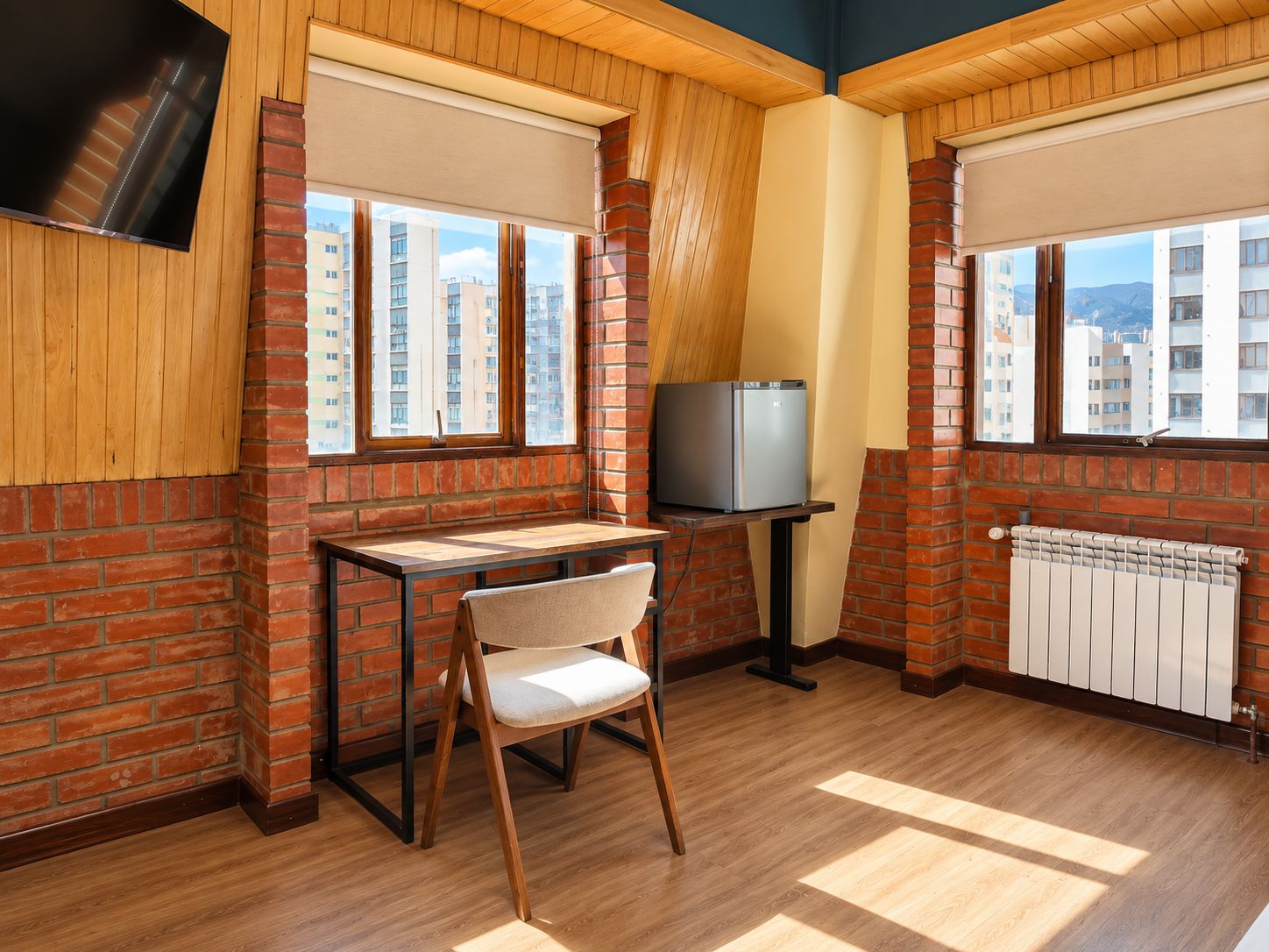 A room with a brick wall, a wooden floor, a television, a small table with a chair, and a radiator.