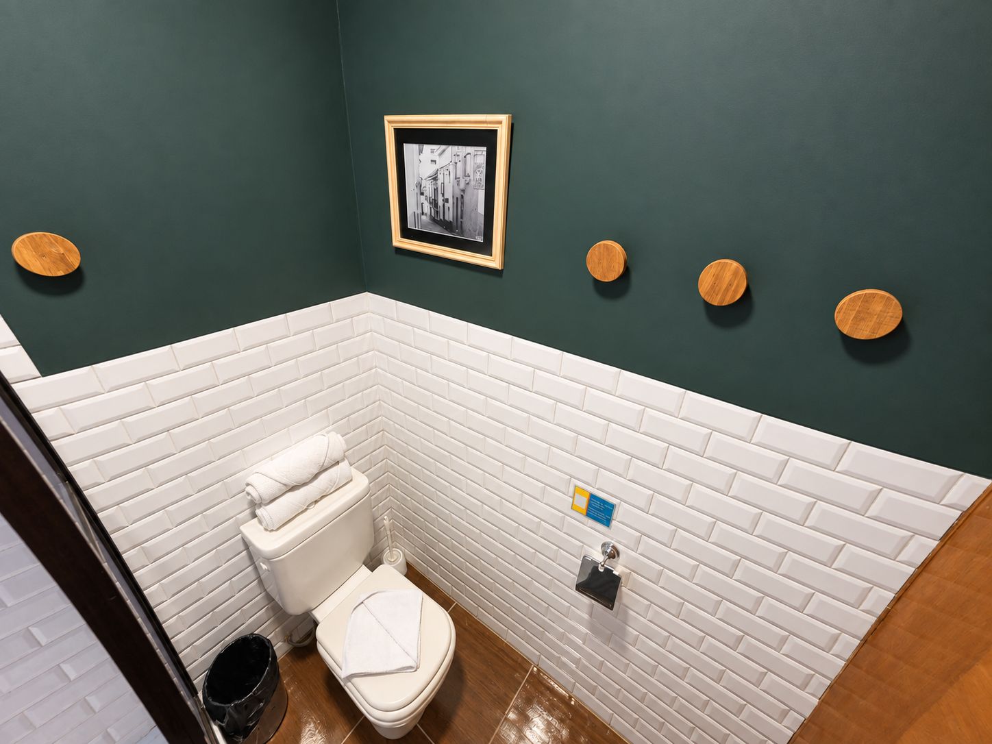 Ein Badezimmer mit weißen Fliesenwänden, einer weißen Toilette, einer schwarzen Mülleimer und Holzdeko.