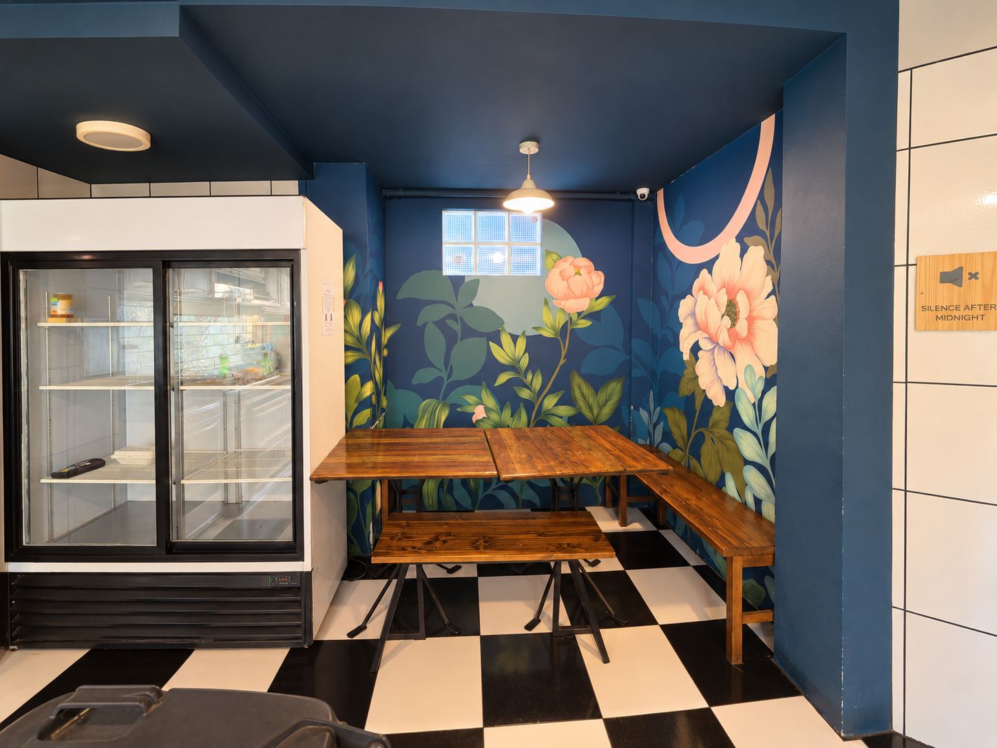 Um quarto com piso xadrez, um banco de madeira, uma geladeira com porta de vidro e um mural floral na parede.