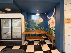 Una habitación con suelo a cuadros, un banco de madera, un refrigerador con puerta de vidrio y un mural floral en la pared.