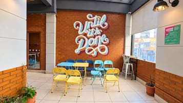 Interior de una cafetería con un letrero que dice 'Until the Dawn', sillas amarillas y azules, y una pared de ladrillo.