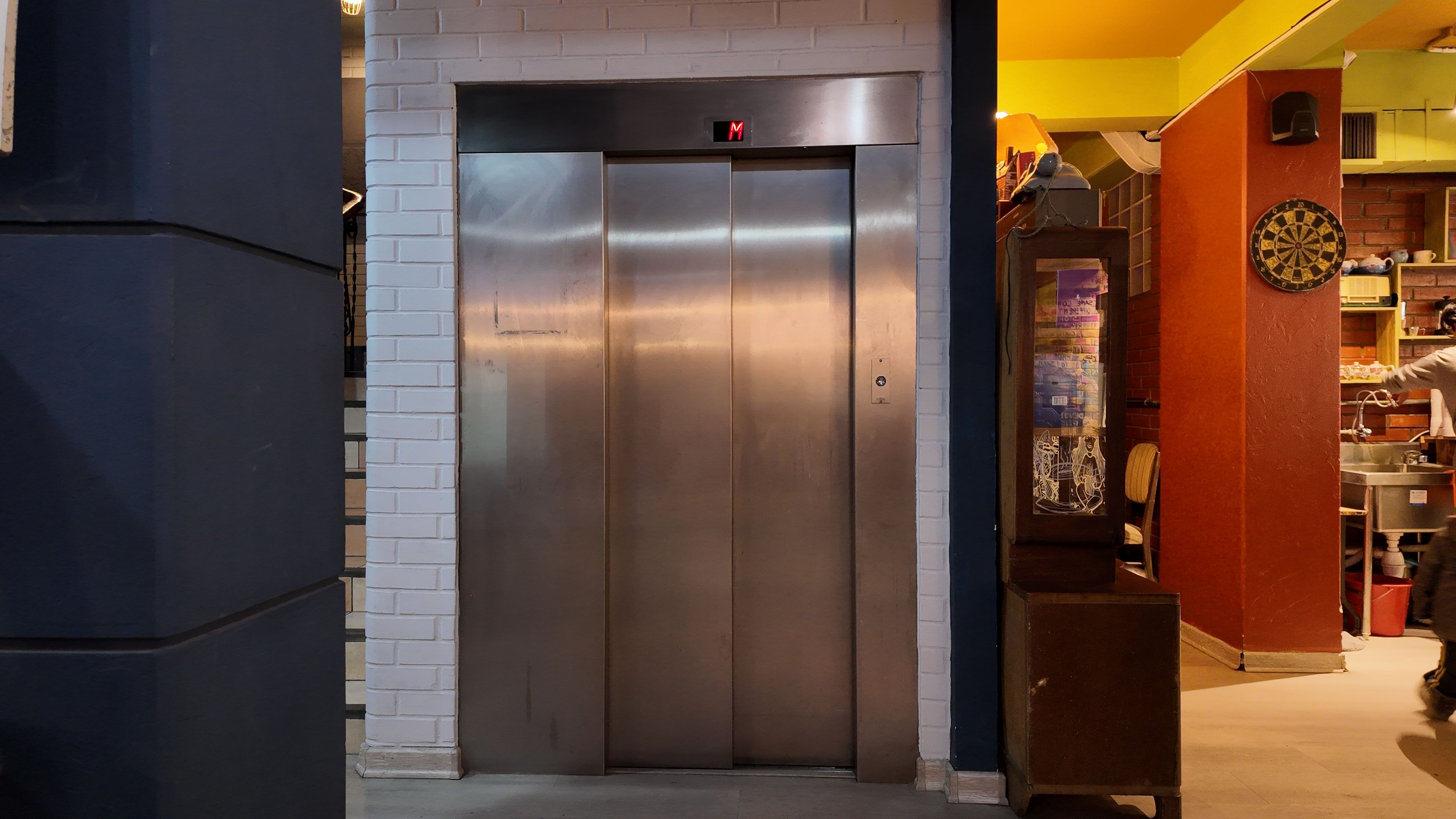 An elevator with a digital display, a brick wall, and a dartboard in the background.