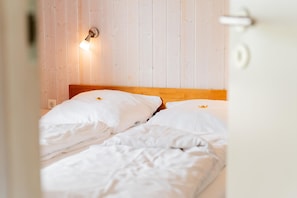 A bed with white bedding and a wooden headboard in a room with wooden panel walls. - Vacation home for 6 people with a sauna and hot tub – fireplace, patio, and large garden (Otterndorf)