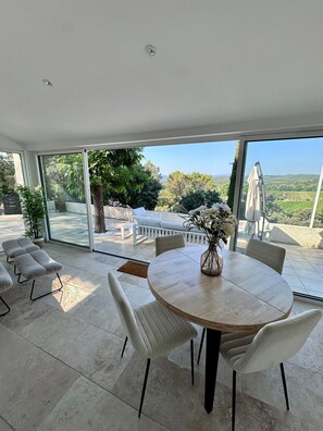 Un espace repas moderne avec une table ronde et des chaises blanches, de grandes fenêtres offrant une vue sur un jardin et un vignoble.