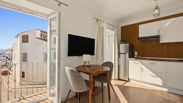 A balcony with a view of buildings, a dining area with a table and chairs, a kitchen with white cabinets and a refrigerator, and a TV mounted on the wall.