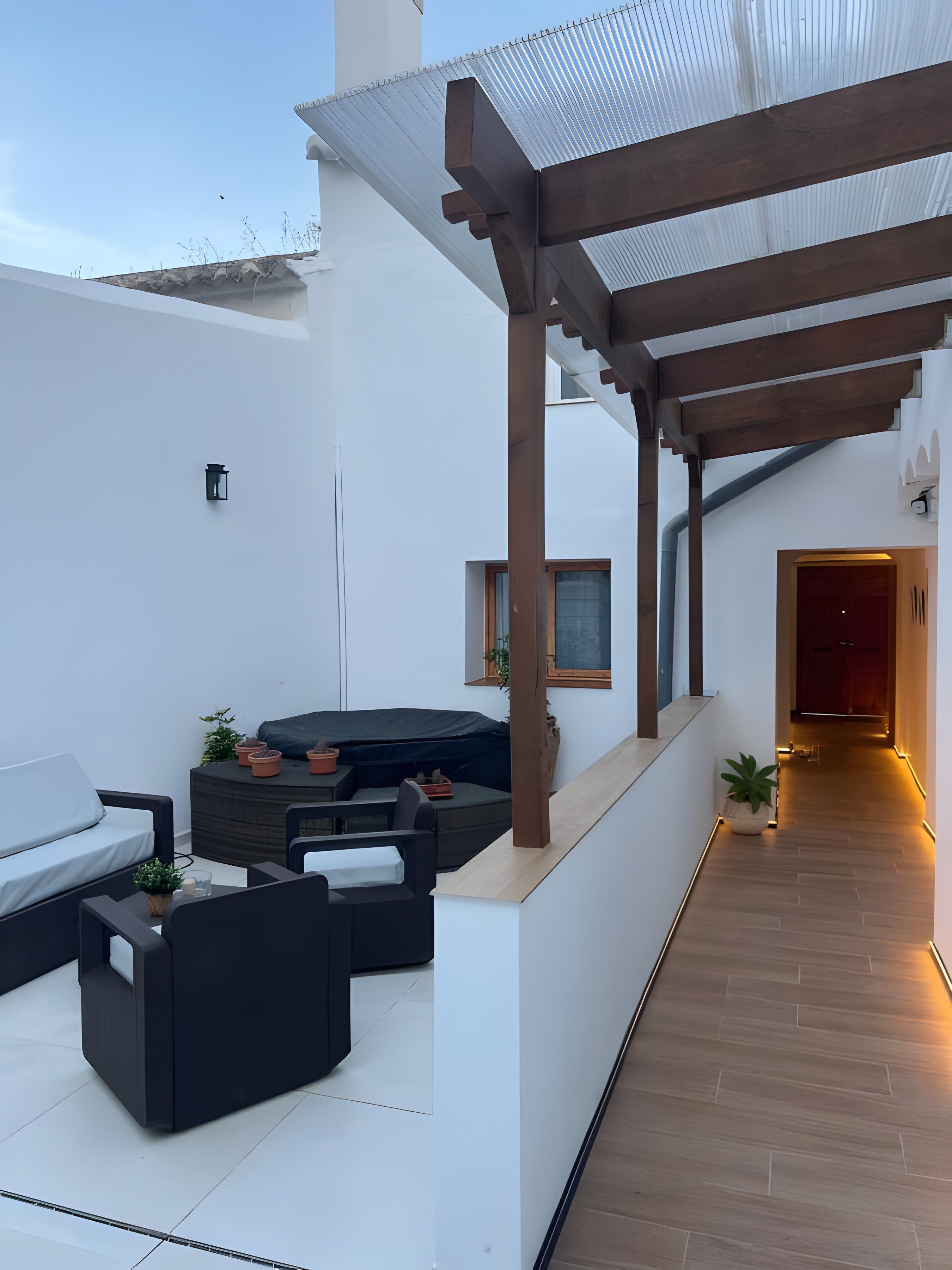 Un patio moderno al aire libre con zona de estar, una pérgola cubierta y suelo de madera que conduce a un pasillo interior.