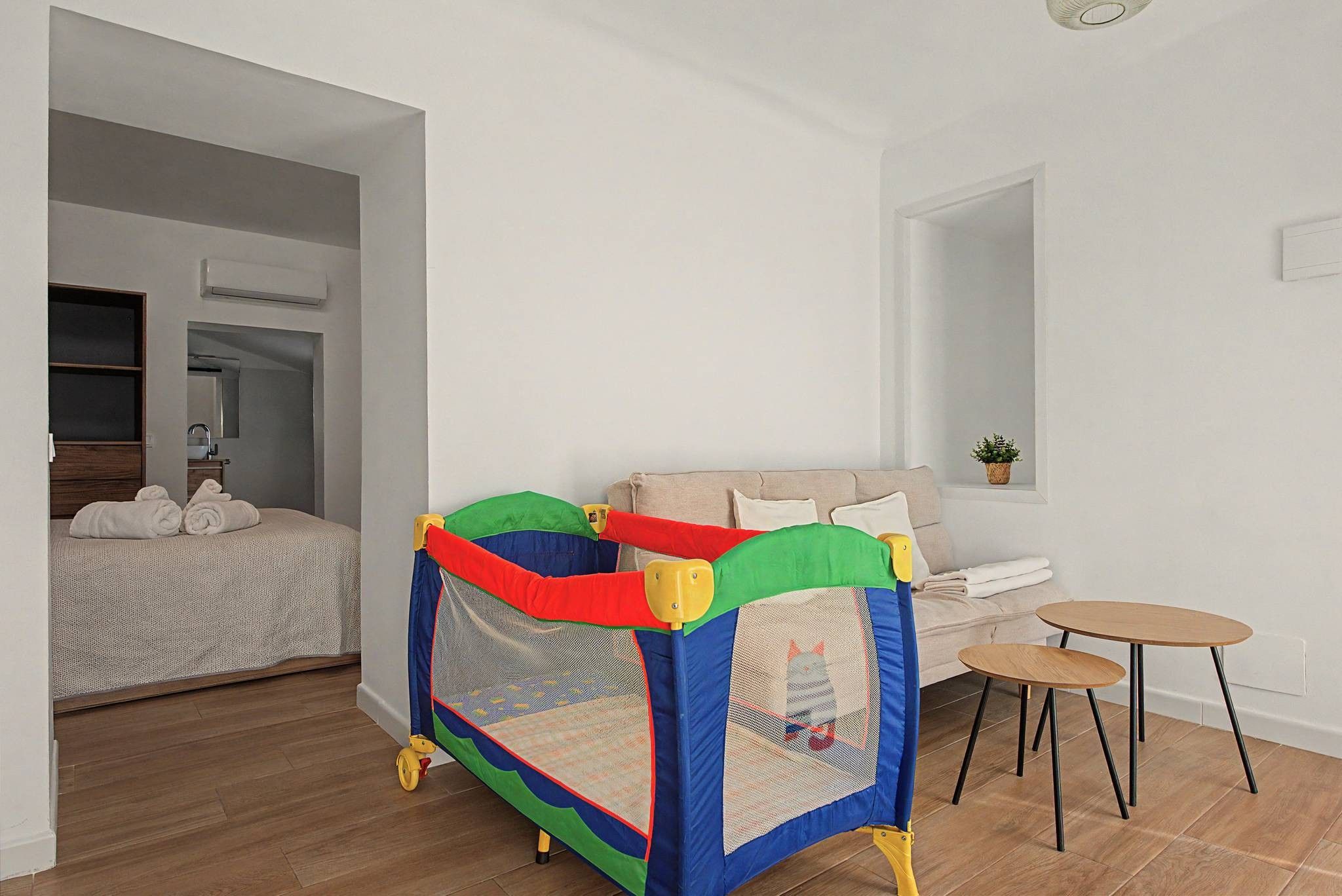 A room with a bed, a small playpen, a sofa, a table, and a potted plant.