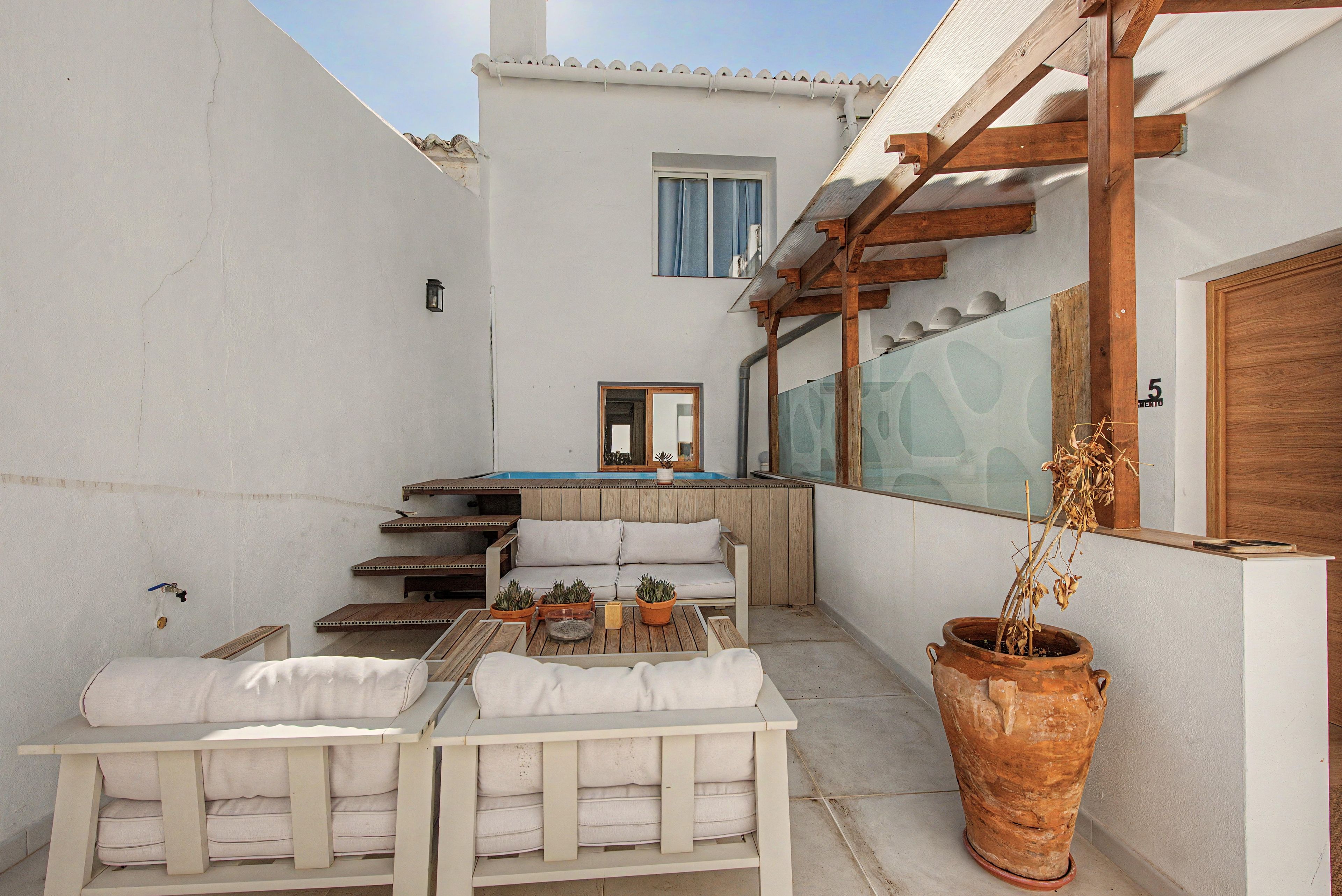Un petit espace extérieur aménagé avec une table et des chaises en bois, un pot de fleurs et une vue sur une piscine et un bâtiment en arrière-plan.