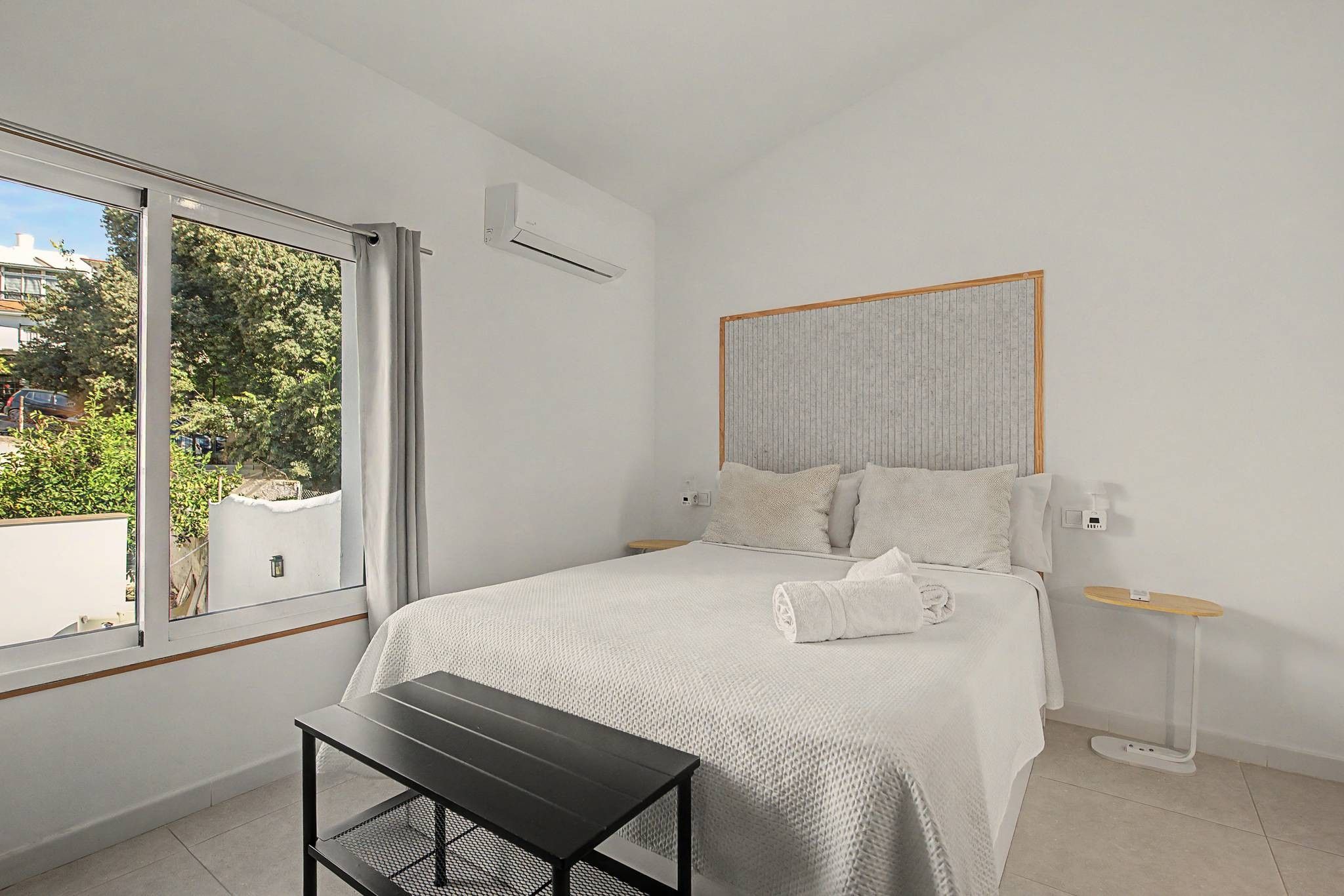 A bedroom with a bed, a bedside table, a nightstand, a window with curtains, and an air conditioning unit.
